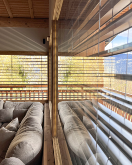 Petit chalet int&eacute;rieur salon avec canap&eacute; confortable face &agrave; une grande baie vitr&eacute;e, vue sur montagne et for&ecirc;t &agrave; travers des stores v&eacute;nitiens en bois.