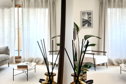Salon lumineux avec canap&eacute;s gris, tables basses ovales et grandes fen&ecirc;tres habill&eacute;es de rideaux en lin, vu en reflet de miroir