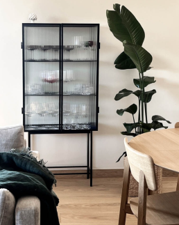 Vitrine noire &agrave; portes en verre cannel&eacute; exposant une collection de verres, dans un salon au parquet ch&ecirc;ne clair avec plante tropicale